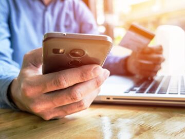 geração Y: homem em frente ao computador olhando celular e segurando cartão de crédito com a outra mão